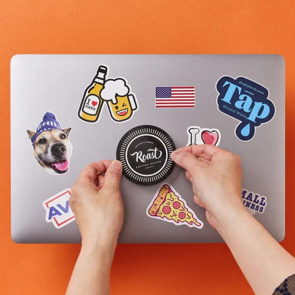 Hands are shown placing a "Roast House Coffee" sticker on a laptop, which is already decorated with various stickers. The stickers include a dog wearing a hat, a beer bottle with "I Beer" text, a smiling beer mug, an American flag, a "Tap Mineral Water" design, an "I " heart, a pizza slice, an "AV" logo, and a "Small Business" label. The background is a solid orange color.